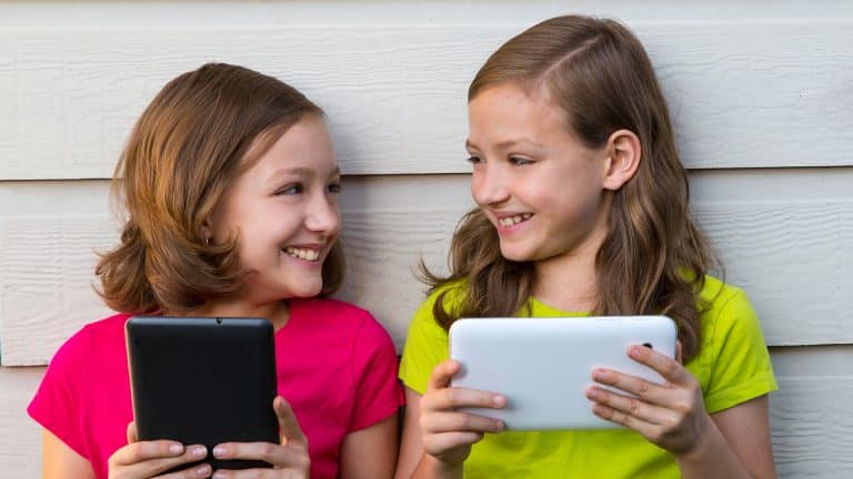 Two happy girls playing with tablet online leaning against a white wall looking each other.